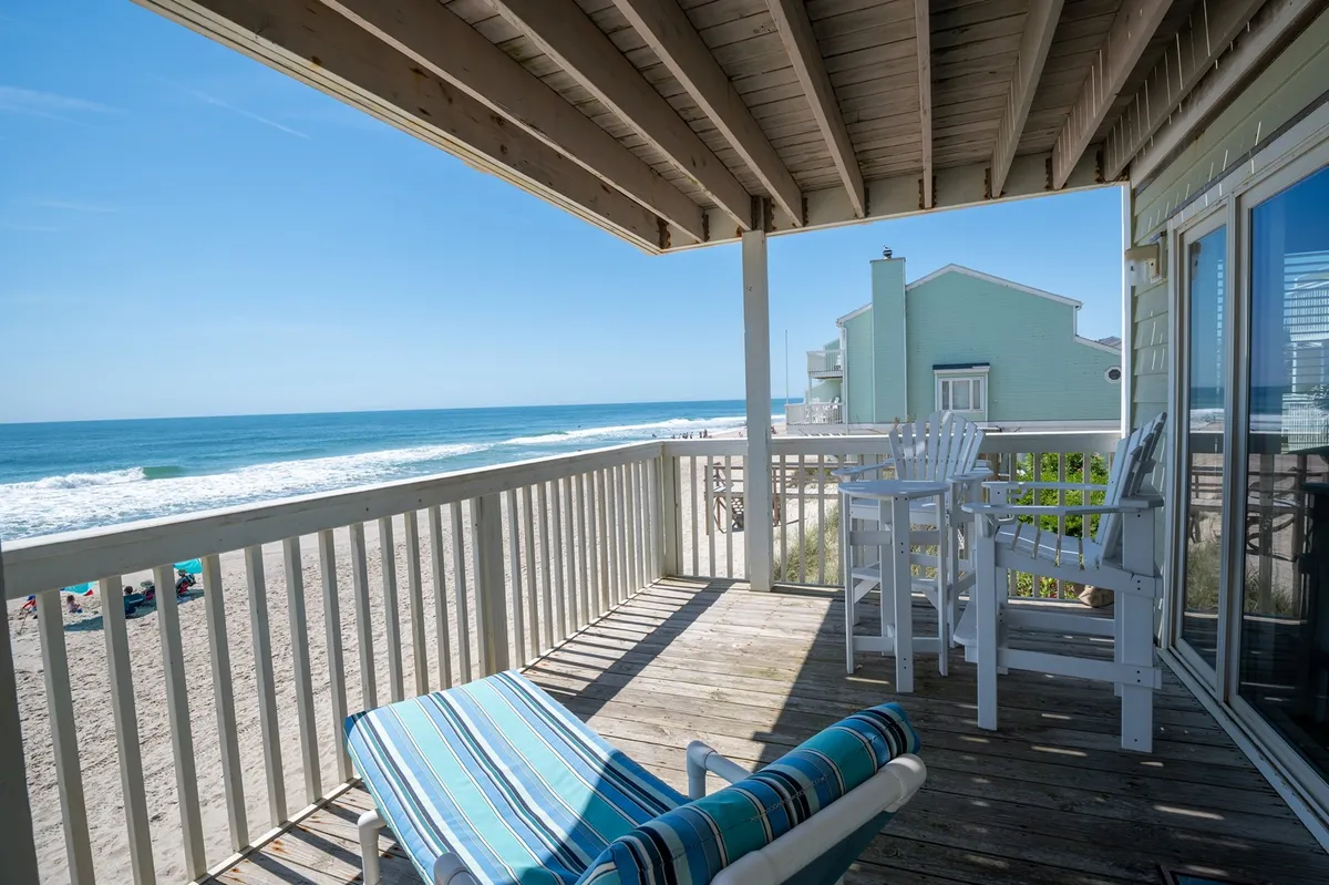 The hero image showcases a tranquil beachfront scene from the condo's private deck, featuring inviting outdoor furniture perfect for relaxing or socializing while overlooking the rolling ocean waves and endless horizon. The view highlights the sunrise sparkling on the water, with pristine sand and blue sky setting a peaceful, dreamy coastal atmosphere.