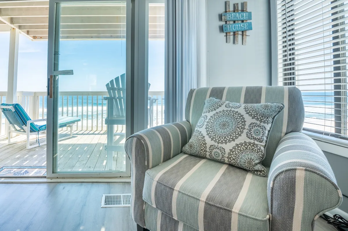 A cozy corner featuring a striped armchair and decorative pillow sits beside a glass door that opens to the balcony, offering a direct view of the beach. A wooden sign reading 'BEACH HOUSE' and serene decor set a relaxed, coastal mood.