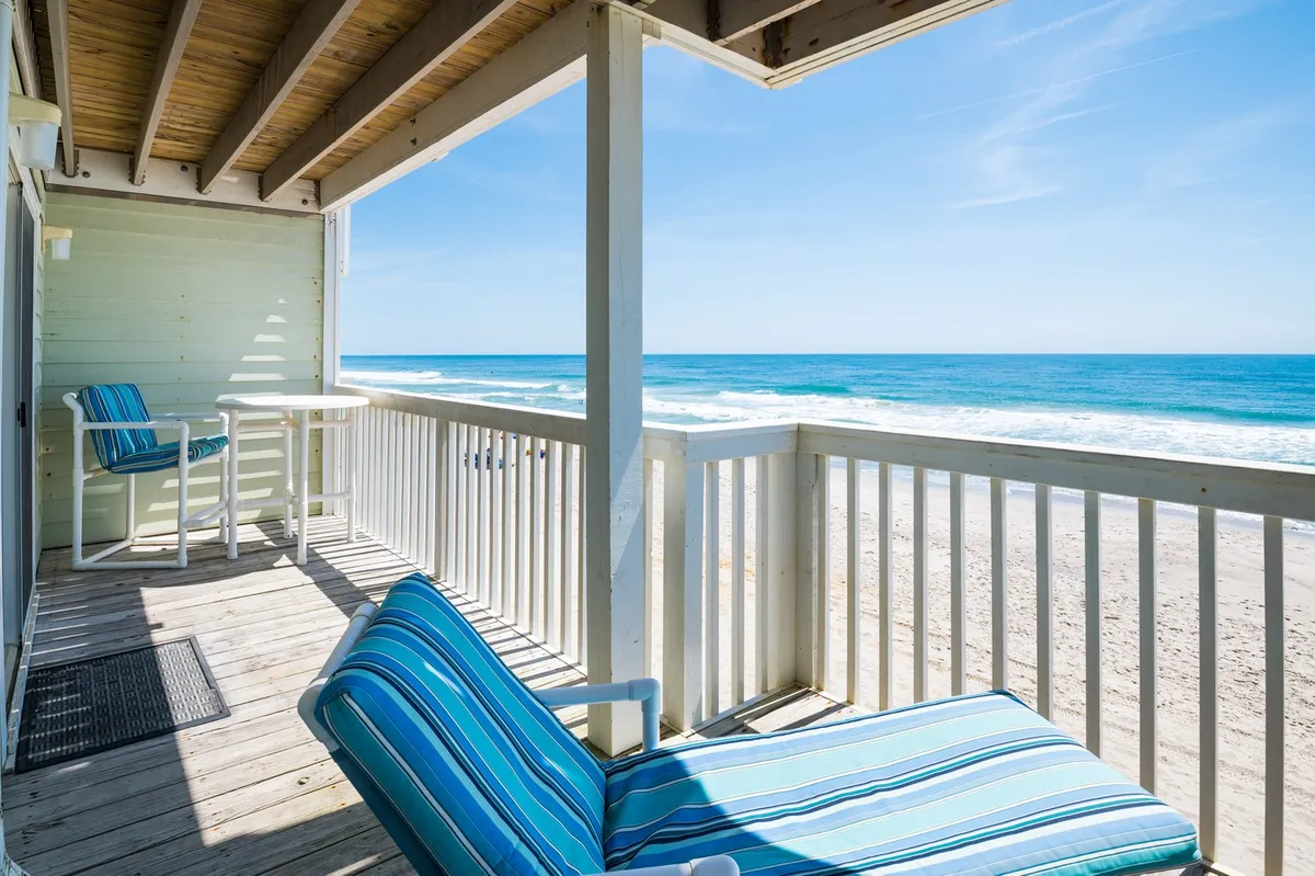 An outdoor deck scene with a striped lounge chair, outdoor table and chairs, all overlooking the sandy beachfront and the Atlantic Ocean beyond. The space is perfect for sunbathing, reading, or socializing with the ocean as your backdrop.