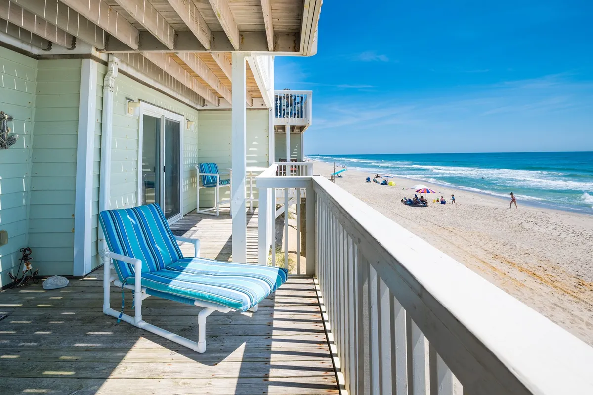 This image captures a tranquil beachfront view from a wooden balcony, with a striped lounge chair positioned to overlook the sand and ocean waves. The inviting scene suggests a direct connection from the property to the beach, with the open sky and water stretching to the horizon.