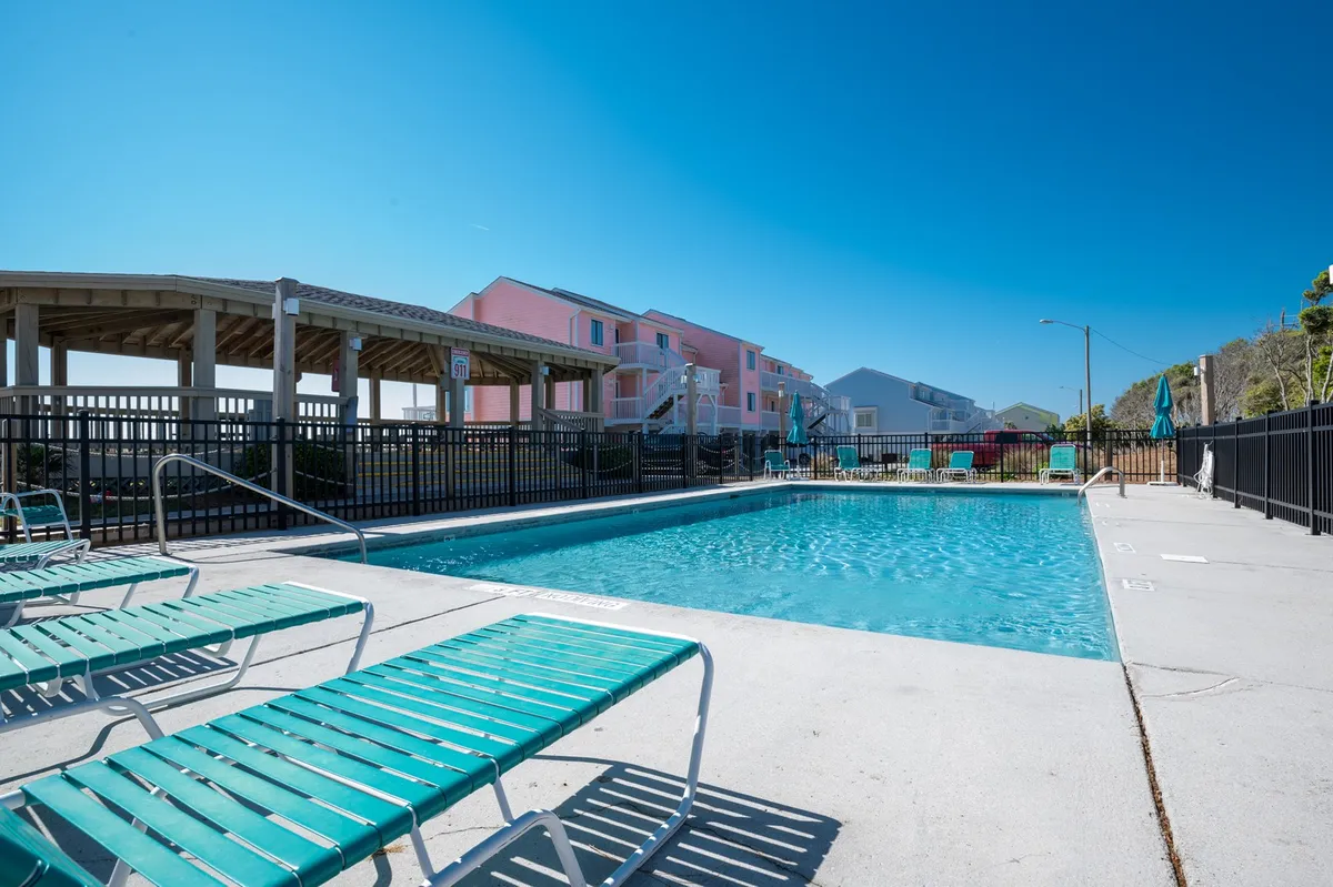 An expansive outdoor swimming pool with lounge chairs and colorful buildings in the background, shaded by poolside umbrellas. The atmosphere is relaxed, inviting for both sunbathing and social gatherings.