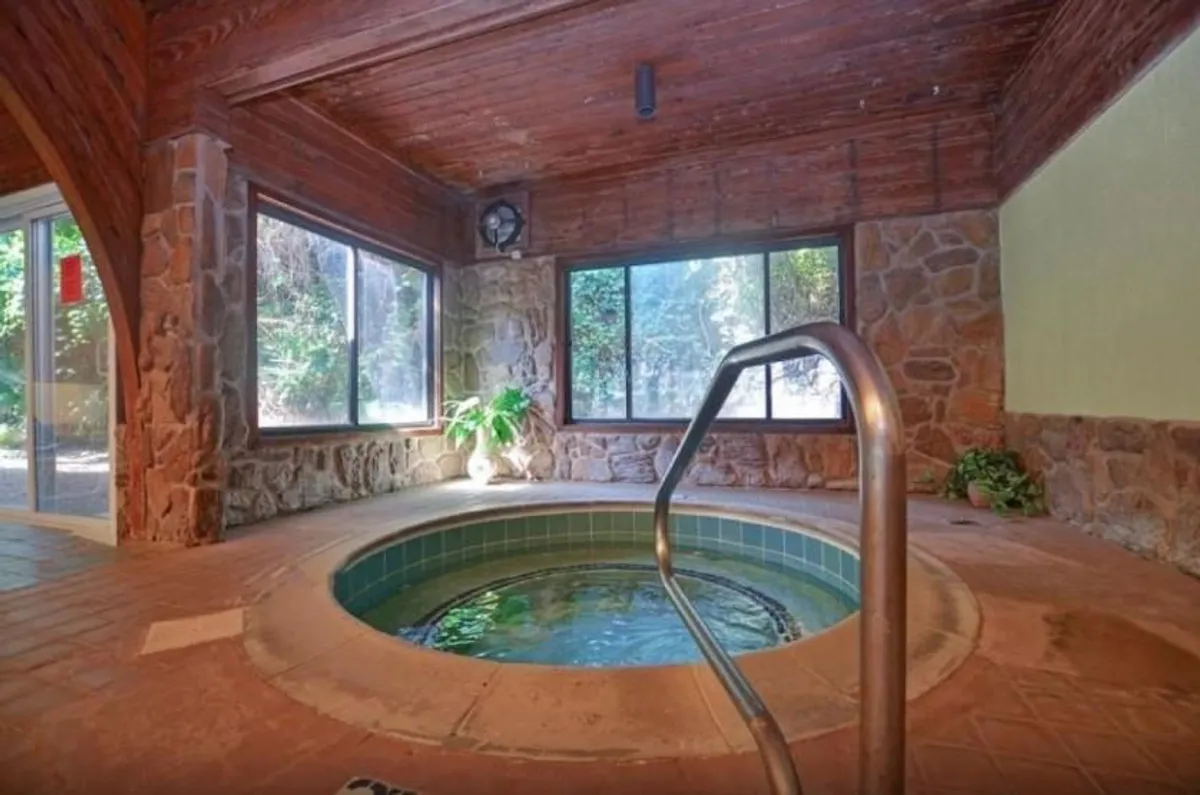An inviting indoor hot tub is set beside large windows, natural stone walls, and a wooden ceiling, allowing natural light to pour in. The scene exudes calm and privacy, with nature views that bring the outside world in.