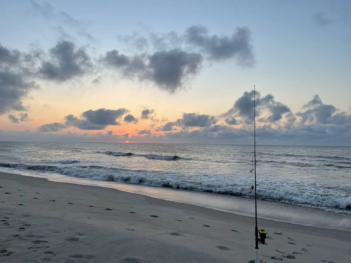 This image captures a tranquil sunset over the ocean, with gentle waves, a fishing rod in the foreground, and soft clouds overhead. The scene suggests a peaceful moment on the beach, ideal for a romantic walk or intimate time together by the water.