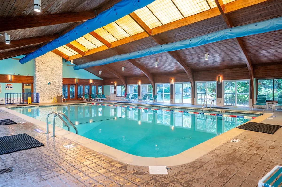 A bright indoor swimming pool area with large windows letting in natural light, lounge chairs lined along the pool, and wooden beams overhead. The space looks ideal for relaxing swims or group activities year-round.