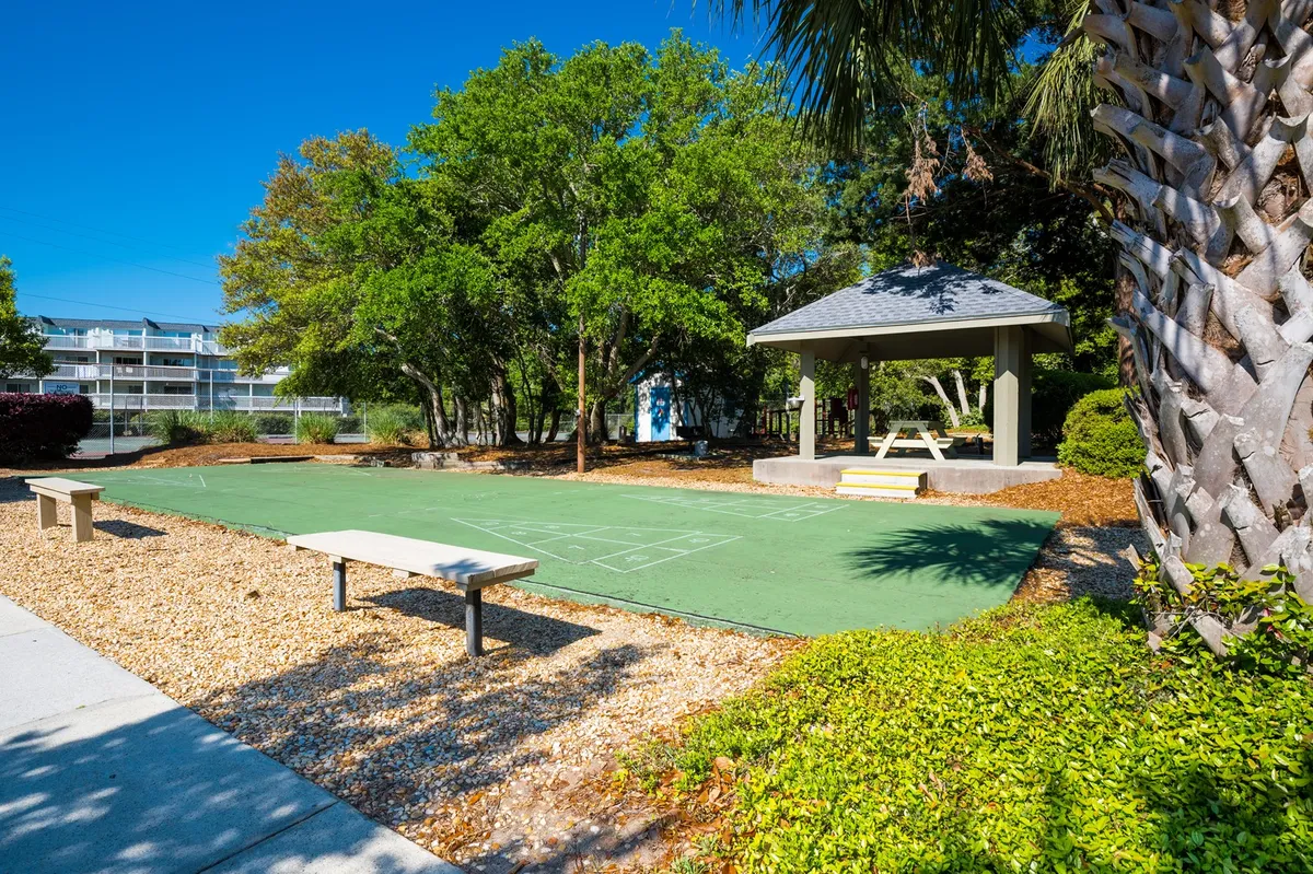This exterior image features a green playing surface surrounded by mature trees, with a charming gazebo and benches offering places to relax. The area is open to the sky and bordered by nature, emphasizing connection to the outdoors and opportunities for play or peaceful reflection.