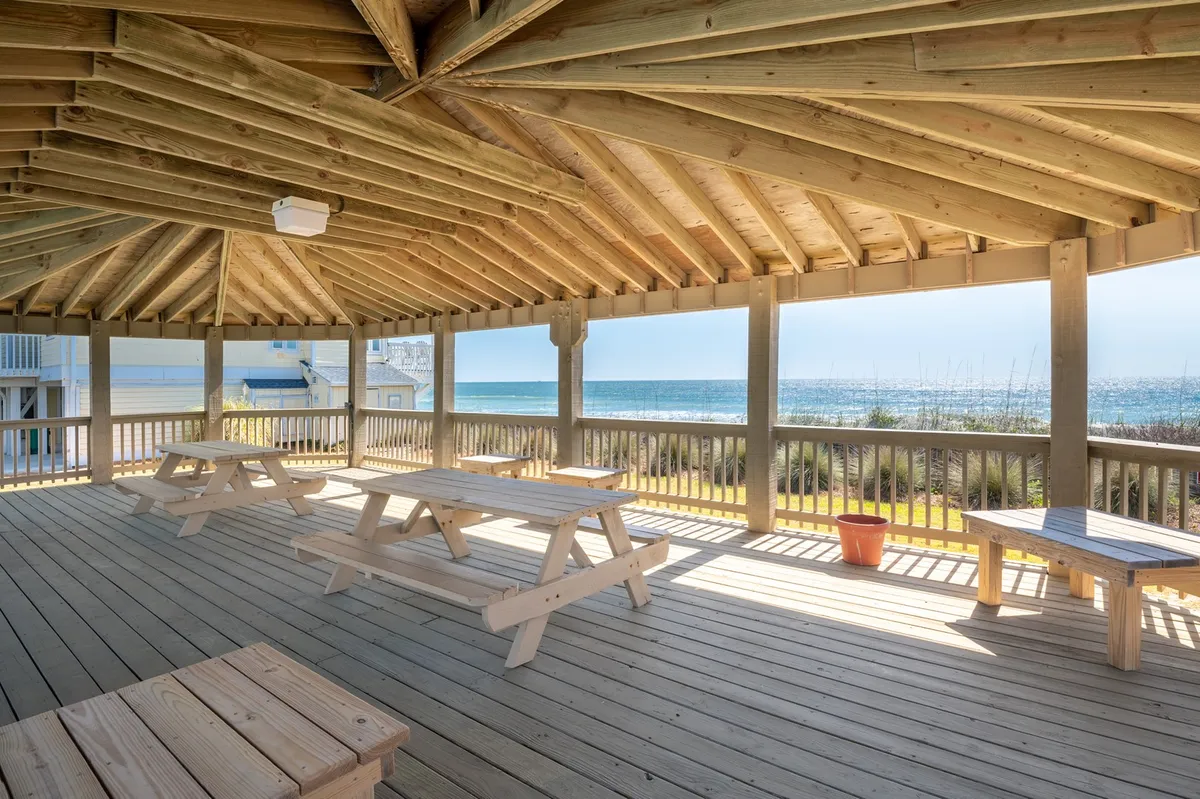 An exterior scene featuring sturdy wooden picnic tables set beneath a shelter, with an expansive ocean view just beyond. The wooden beams add a coastal vibe, and the area is spacious enough for group gatherings, picnics, or outdoor meals. The natural light and cheerful atmosphere make it an inviting spot for multi-generational travelers to gather.