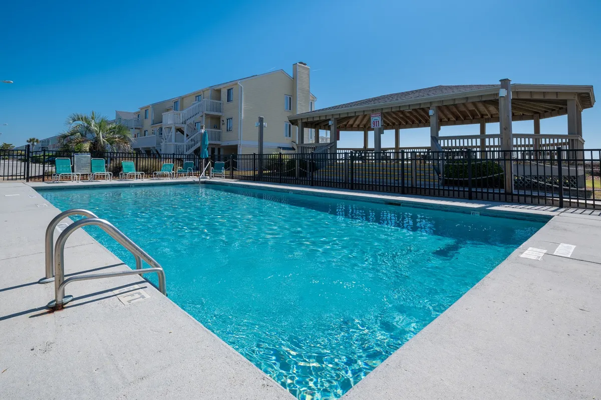 A spacious outdoor swimming pool area bordered by lounge chairs, a gazebo, and lush greenery. The pool sparkles in the sunlight, offering ample space for family swims, sunbathing, and relaxation after a day at the beach.