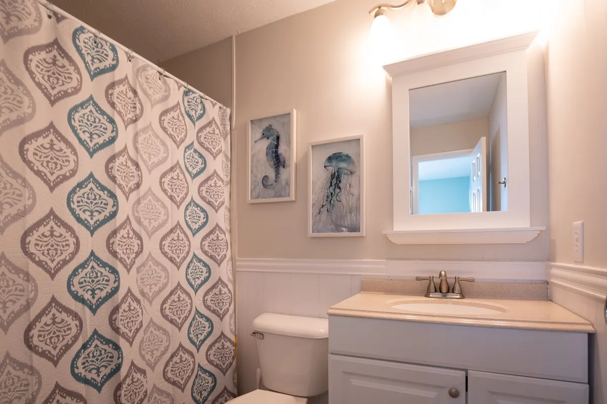 This bathroom features a stylish vanity area with a sink, a mirror, and a shower adorned with an intricately patterned curtain. Subtle coastal-themed artwork decorates the wall, and the space is organized and fresh, conveying a sense of cleanliness and modern comfort.