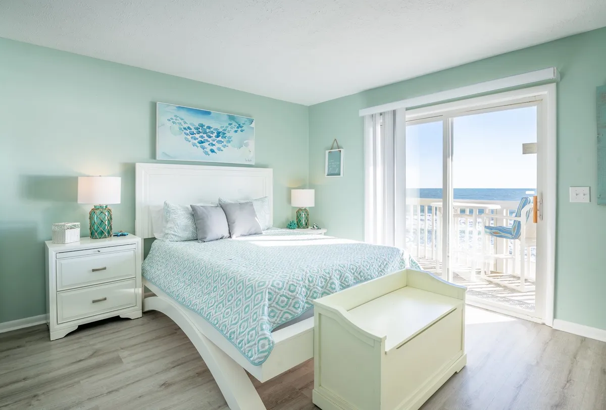 This image showcases a well-appointed bedroom featuring a queen bed with crisp linens, a nightstand, decorative artwork, and expansive windows revealing a stunning ocean view. The tranquil ambiance is enhanced by soft lighting and serene decor, inviting guests to relax and unwind. The room also has direct access to an oceanfront balcony, emphasizing privacy and direct connection to the coastal scenery.