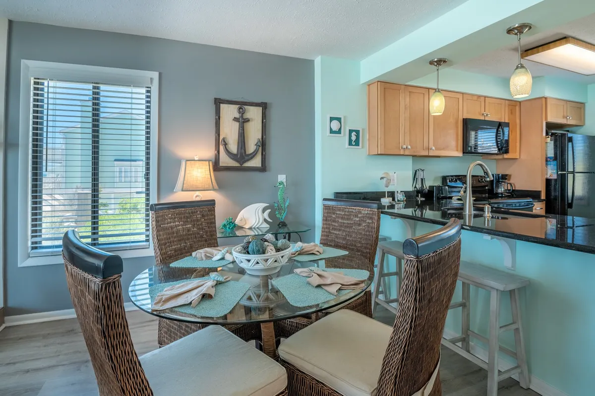 The image shows a round dining table with nautical anchor wall decor and a view into the kitchen area. It creates a welcoming, cheerful atmosphere for dining and gathering, while also giving another angle of the well-equipped kitchen.