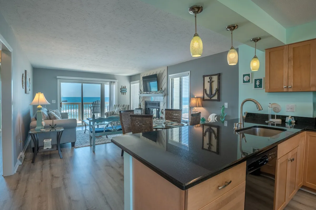This image captures an open-plan interior featuring the kitchen island, dining table, and living area, all oriented towards expansive windows with ocean views. The living area is designer-furnished, with a large TV and fireplace, and the decor resonates with coastal themes (anchors, seashells).